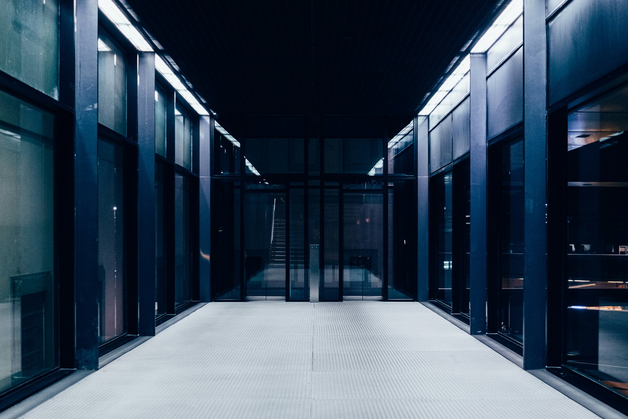 Modern, symmetrical corridor with glass walls and dark structural columns leading to a central doorway, conveying precision, structure, and controlled execution.