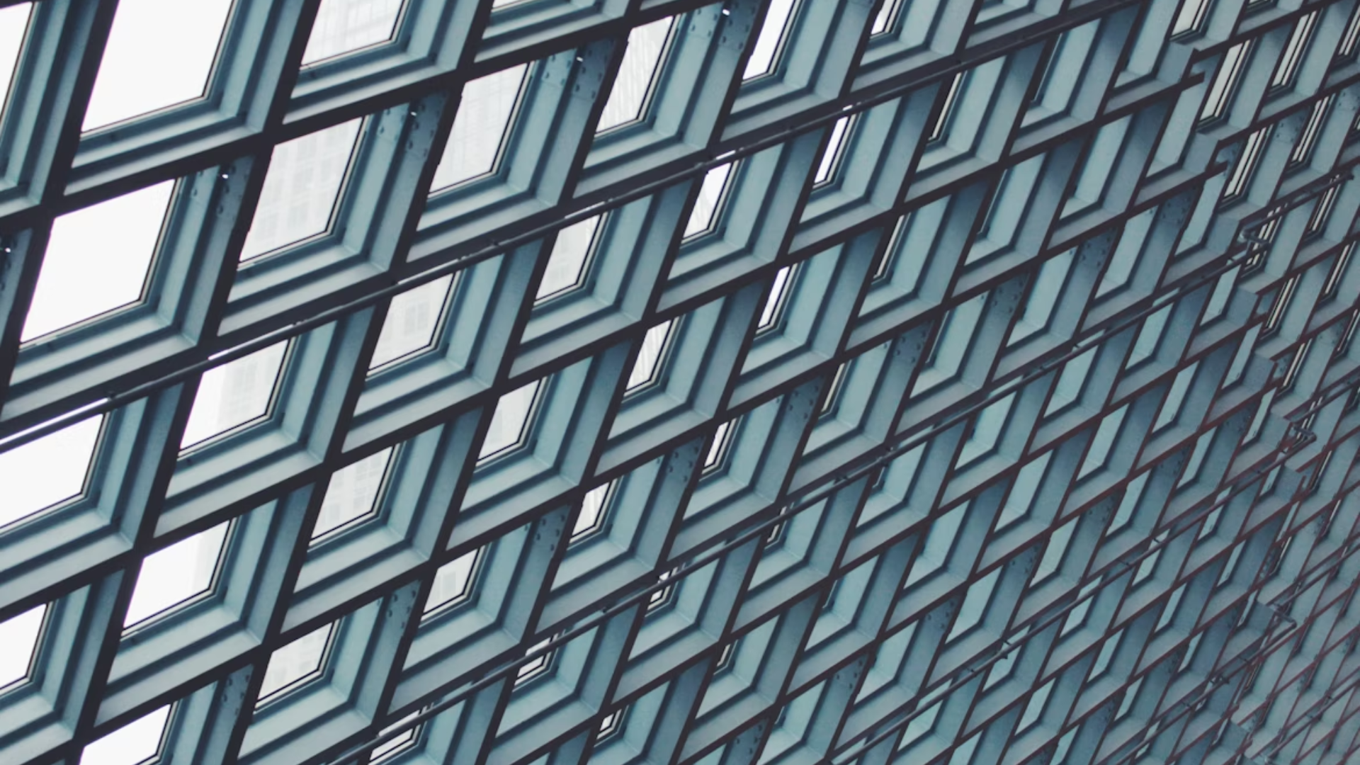 Diagonal view of a modern glass building facade with repeating geometric window frames in a grid pattern, creating a layered, architectural texture.